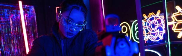 armed asian woman in sunglasses aiming with gun near neon sign, banner - Foto, Imagen