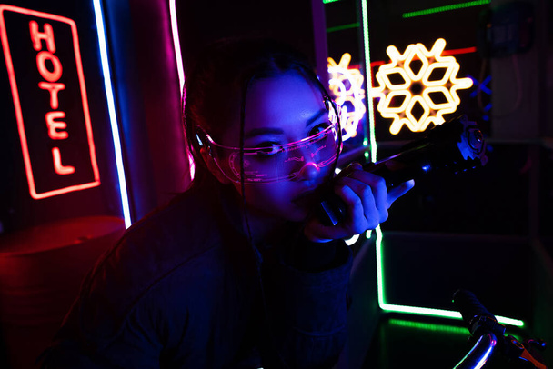 armed asian woman in sunglasses holding gun near neon hotel sign  - Zdjęcie, obraz