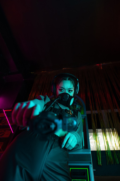 Tiefansicht einer bewaffneten asiatischen Frau mit Gasmaske und drahtlosem Kopfhörer, die mit der Waffe in die Kamera schaut  - Foto, Bild