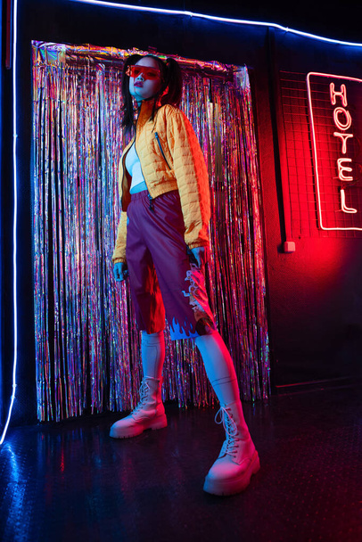 full length of young asian woman in sunglasses looking at camera near red neon hotel sign  - Zdjęcie, obraz
