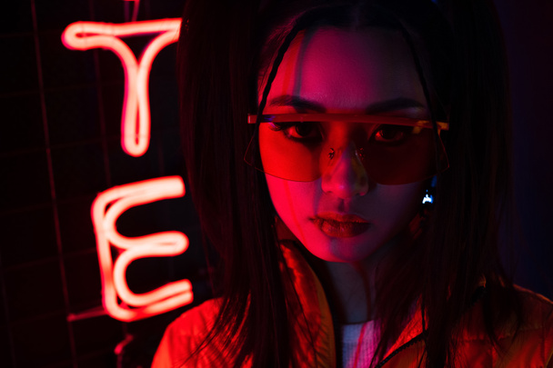 Beleuchtung einer jungen Asiatin mit Sonnenbrille, die in der Nähe einer roten Leuchtreklame in die Kamera blickt  - Foto, Bild