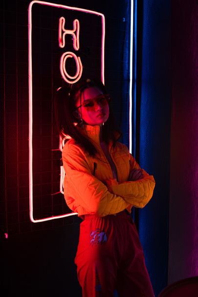 young asian woman in sunglasses standing with crossed arms near red neon hotel sign  - Photo, Image