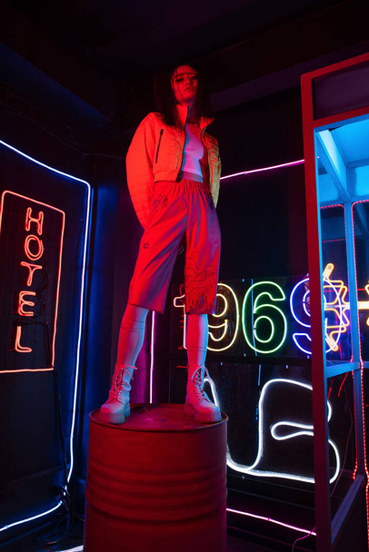 full length of asian woman in sunglasses looking at camera while posing near red neon hotel sign  - Zdjęcie, obraz