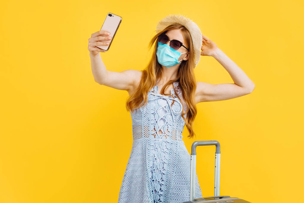 Young woman in summer hat and medical mask making video call from smartphone using online conference app while standing with suitcase on yellow background - Foto, imagen