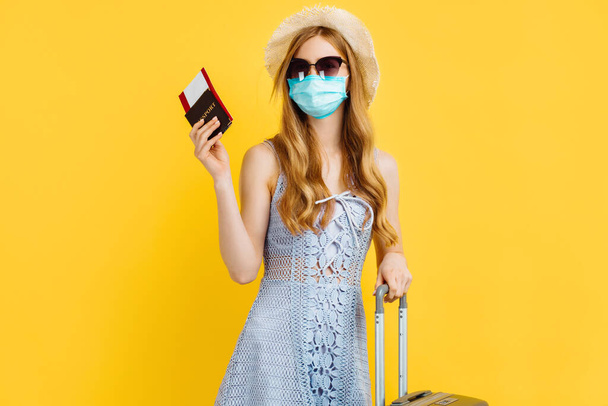 Cheerful young woman in protective mask dressed in summer clothes holding passport with tickets while standing with suitcase on yellow background - Fotó, kép