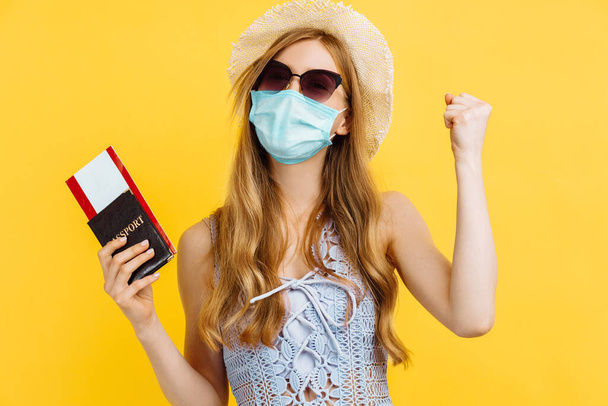 Cheerful young woman in protective mask dressed in summer clothes holding passport with tickets while standing on isolated yellow background - 写真・画像