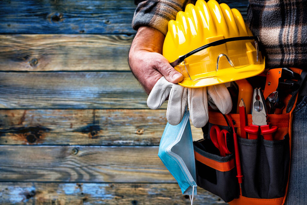 Elektriker auf hellblauem Holzgrund; hält Helm, Handschuhe, Schutzbrille und Operationsmaske, um die Ausbreitung des Coronavirus zu verhindern. Bauindustrie, Arbeitssicherheit. - Foto, Bild