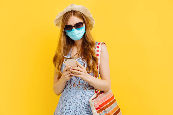 Young happy woman in medical mask, in summer dress and hat uses mobile phone isolated over yellow background, Summer concept, vacation - Photo, Image