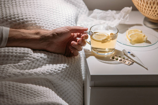 Zieke man ligt in bed onder de deken en houdt een kop warme thee met citroen vast, close-up. Man die warme drank drinkt om beter te worden van griep, koorts en virus. Griepseizoen.  - Foto, afbeelding