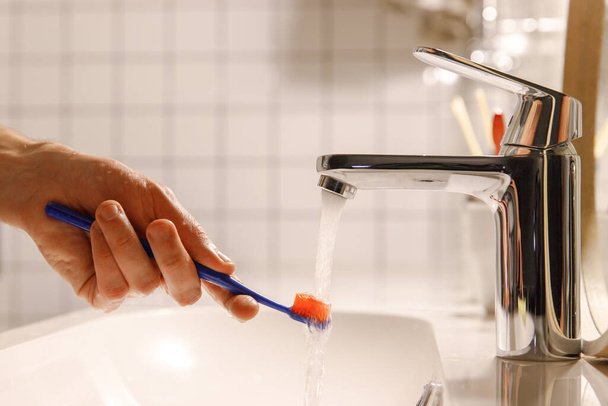 Hombre limpiando su cepillo de dientes bajo el agua corriente en el baño, limpiándolo, enfoque suave. Procedimientos para dormir, higiene bucal / odontológica. - Foto, Imagen