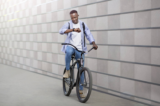 Ευτυχισμένος Αφροαμερικάνος με ακουστικά που ακούει μουσική και ελέγχει την ώρα στο SmartWatch κατά τη διάρκεια της ποδηλασίας έξω - Φωτογραφία, εικόνα