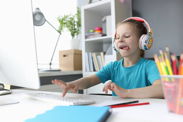 Little girl in headphones sitting at table and typing on computer keyboard - 写真・画像