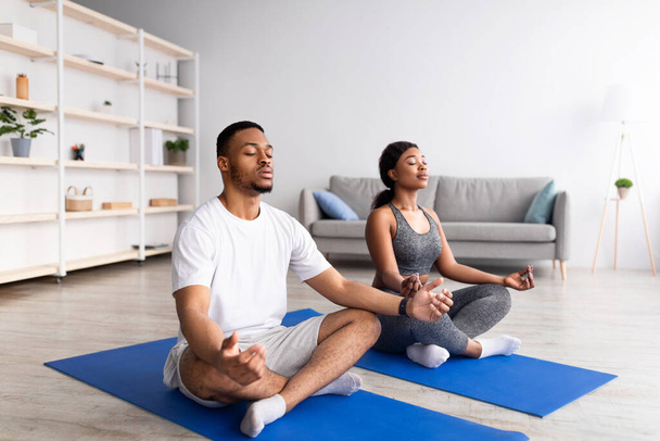 Young black couple meditating with closed eyes, sitting on sports mats in lotus pose at home, full length - Foto, afbeelding
