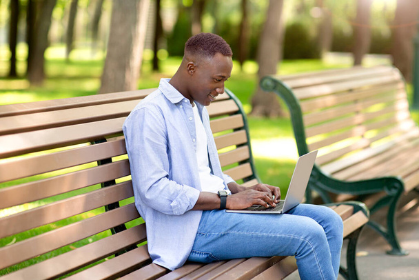 Portrait of cool black guy using laptop on bench at park, working remotely, communicating on webcam - Photo, Image