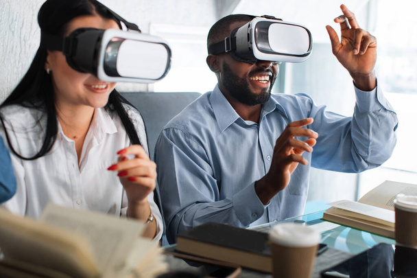 Digital 3d virtual technology. Education with modern gadgets and technologies. Happy smiling positive two friends, Asian girl and African man, moving hands while trying VR goggles. - Foto, afbeelding