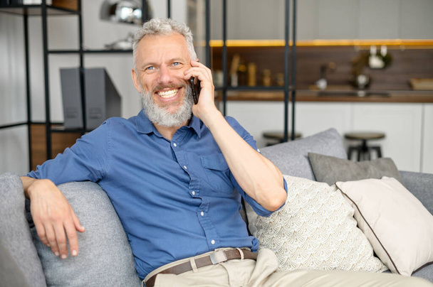 Senior bearded guy sitting down on the comfortable couch and talking on the smartphone - Foto, imagen