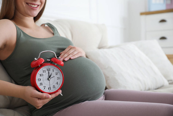 Young pregnant woman holding alarm clock near her belly at home, closeup. Time to give birth - Fotó, kép