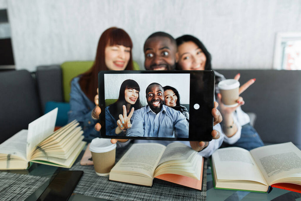 Group of three young multiethnic friends, sitting at the table in cafe, reading books, taking selfie using digital tablet pc, laughing, gesturing. Focus on tablet computer. - 写真・画像