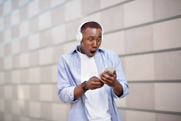Hombre afroamericano sorprendido en auriculares mirando la pantalla del teléfono inteligente en la calle de la ciudad - Foto, imagen