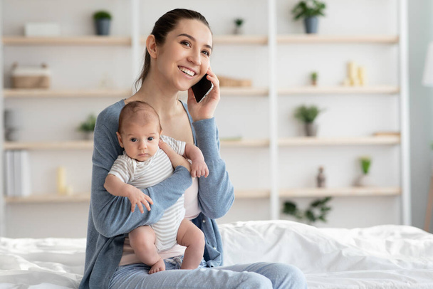 若い母親のライフスタイルの概念。笑顔のお母さんが電話で話し赤ちゃんを抱いて家でノートパソコンを使う. - 写真・画像