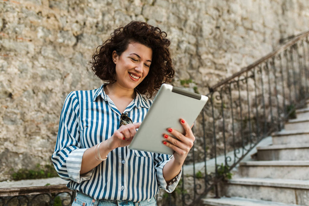 Attraktive junge Frau mit lockigem Haar bedient ihr Touchscreen-Tablet, während sie draußen steht - Foto, Bild