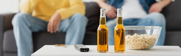 bottles of beer, smartphone, tv remote and popcorn near friends sitting on blurred background, banner - Photo, Image