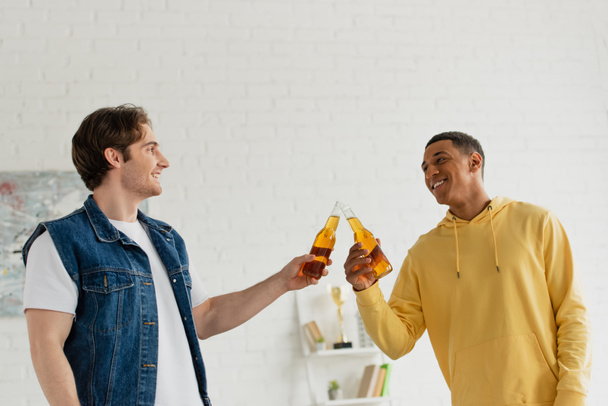 happy young interracial friends clinking with bottles of beer in modern loft - Photo, Image
