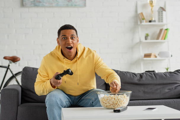 KYIV, UKRAINE - MARCH 22, 2021: excited african american man sitting with joystick on couch in living room - Foto, afbeelding