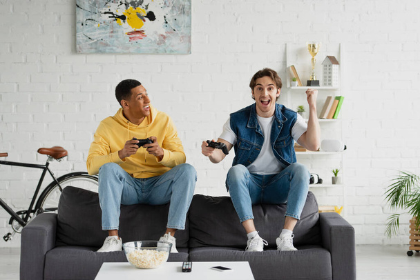 KYIV, UKRAINE - MARCH 22, 2021: young hipster playing video game with african american friend in modern loft - Photo, Image