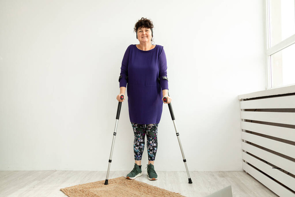 Mujer de edad se levanta sobre fondo blanco en muletas en la oficina de fisioterapeuta. Concepto de recuperación y rehabilitación de traumas - Foto, imagen