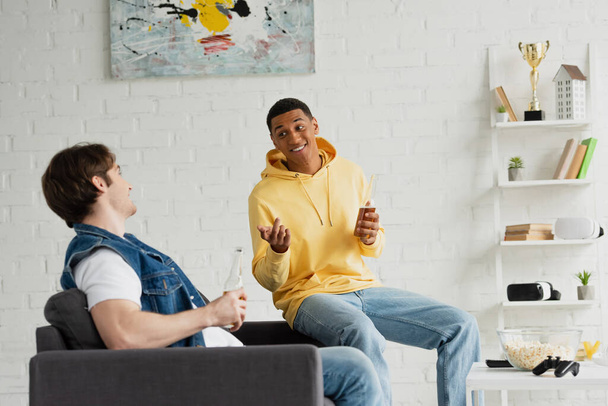 KYIV, UCRANIA - 22 de marzo de 2021: hombres interracial conversando amistosamente y sentados en el sofá con botellas de cerveza en la sala de estar  - Foto, imagen