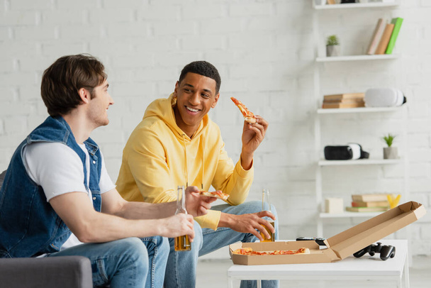 KYIV, UKRAINE - MARCH 22, 2021: african american smiling man eating pizza and drink beer with friend on couch in living room - Фото, изображение