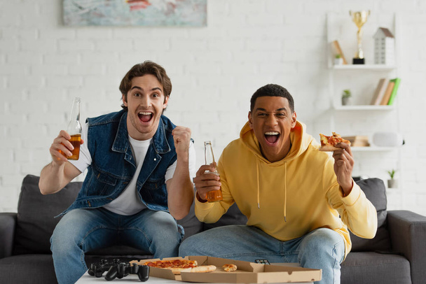 KYIV, UKRAINE - MARCH 22, 2021: interracial friends sitting on couch with bottles of beer and pizza  - Foto, afbeelding