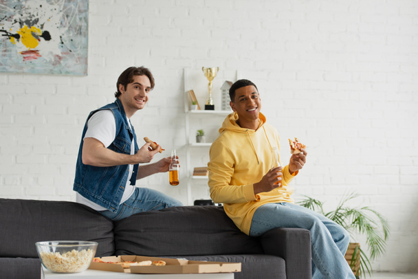 young interracial friends enjoying pizza and beer on couch in modern living room - 写真・画像