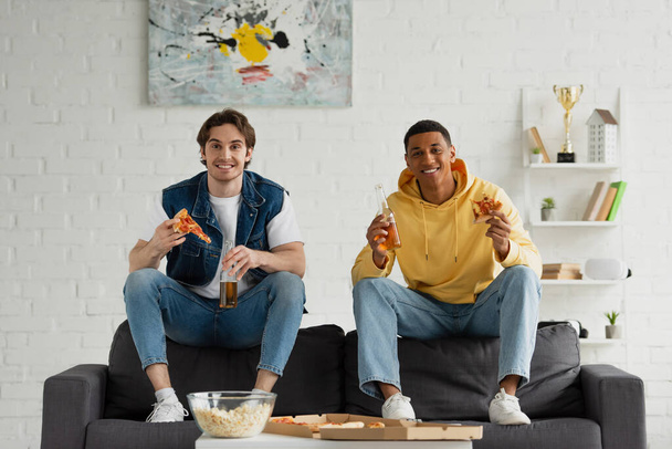 happy interracial friends enjoying pizza slices, popcorn and beer on couch in modern living room - Photo, Image