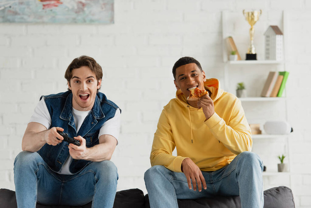 KYIV, UKRAINE - MARCH 22, 2021: interracial friends playing video game and enjoying pizza on couch in modern living room - Zdjęcie, obraz