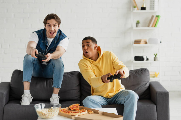 KYIV, UKRAINE - MARCH 22, 2021: Různé přátelé emocionálně hrají videohry s joysticky a užívají si pizzu na gauči v moderním obývacím pokoji - Fotografie, Obrázek