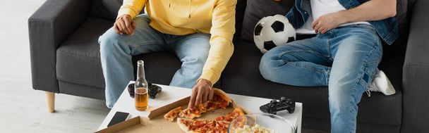 KIEW, UKRAINE - 22. MÄRZ 2021: Teilansicht von Männern in Jeans, die auf einer Couch neben einem Tisch mit Pizza, Bierflasche, Popcorn, Steuerknüppel und Ball sitzen, Banner - Foto, Bild