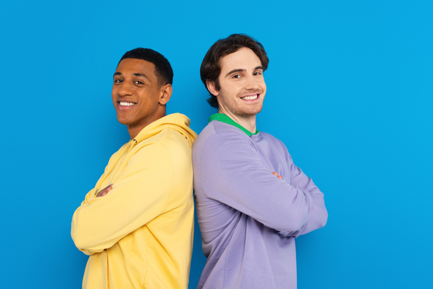 glimlachen interraciale vrienden in rug naar rug pose geïsoleerd op blauw - Foto, afbeelding