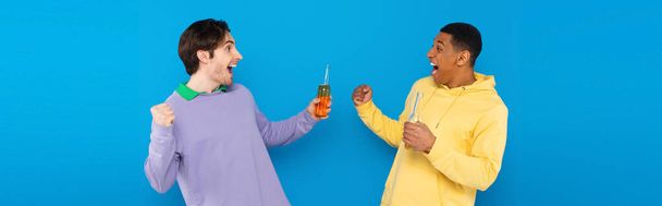 happy interracial friends celebrating with bottles of beer isolated on blue, banner - Photo, Image