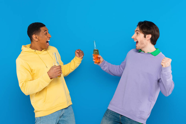 szczęśliwy międzyrasowy przyjaciele uśmiechając się i świętując z butelki piwa odizolowany na niebiesko - Zdjęcie, obraz