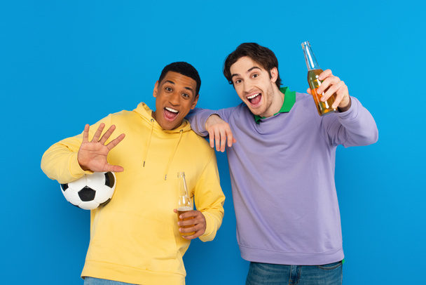 aficionados al deporte interracial emocionados celebración de fútbol y botella de cerveza aislada en azul - Foto, imagen