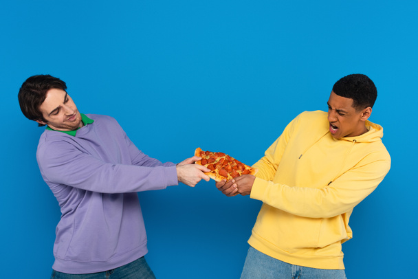 międzyrasowi przyjaciele emocjonalnie dzielą się jednym kawałkiem pizzy izolowanym na niebiesko - Zdjęcie, obraz