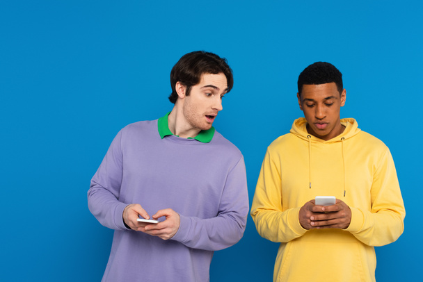 curioso hipster sbirciare smartphone di afro americano amico isolato su blu - Foto, immagini