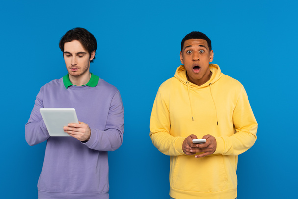 hipsters interracial sosteniendo gadgets en manos aisladas en azul - Foto, imagen