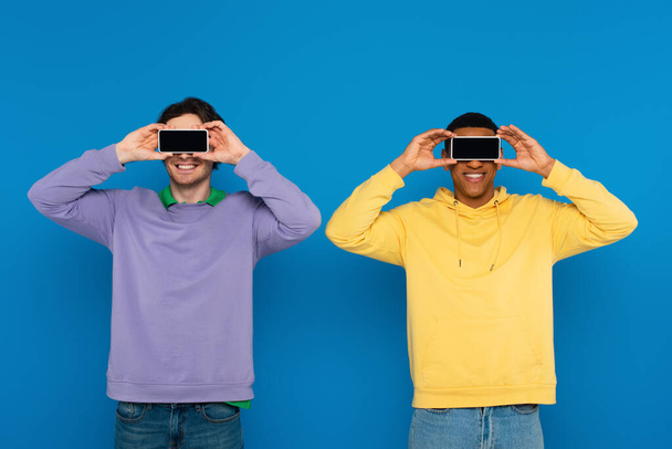 Irklar arası hipsterlar mavi üzerine izole edilmiş boş ekranlı akıllı telefonları olan gözleri kapatıyorlar. - Fotoğraf, Görsel