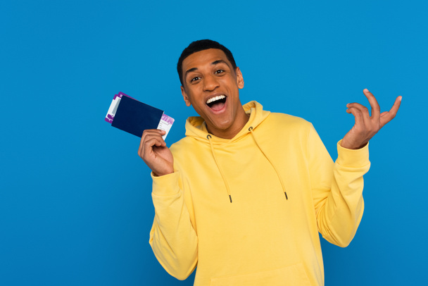 heureux homme afro-américain titulaire d'un passeport avec des billets d'avion dans les mains isolé sur bleu - Photo, image