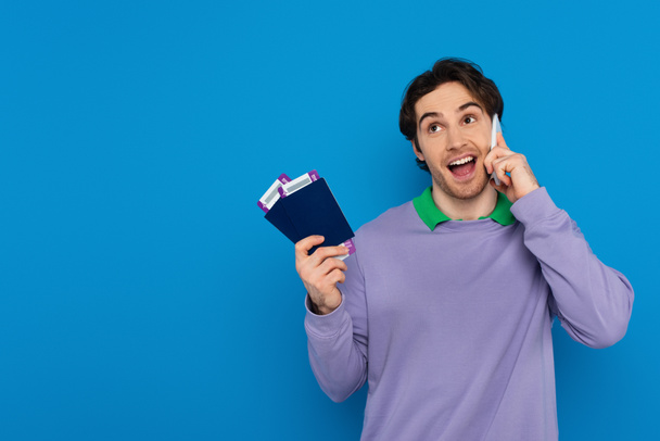 幸せな若い男が手に航空券を持ってパスポートを保持し、青で隔離された携帯電話で話す - 写真・画像