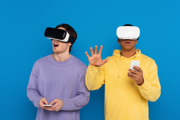 excited interracial hipsters playing video game with virtual reality headsets isolated on blue - Фото, зображення