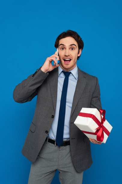 junger Mann im modischen Anzug hält Geschenkbox mit roter Schleife und spricht auf Handy isoliert auf blauem Grund - Foto, Bild